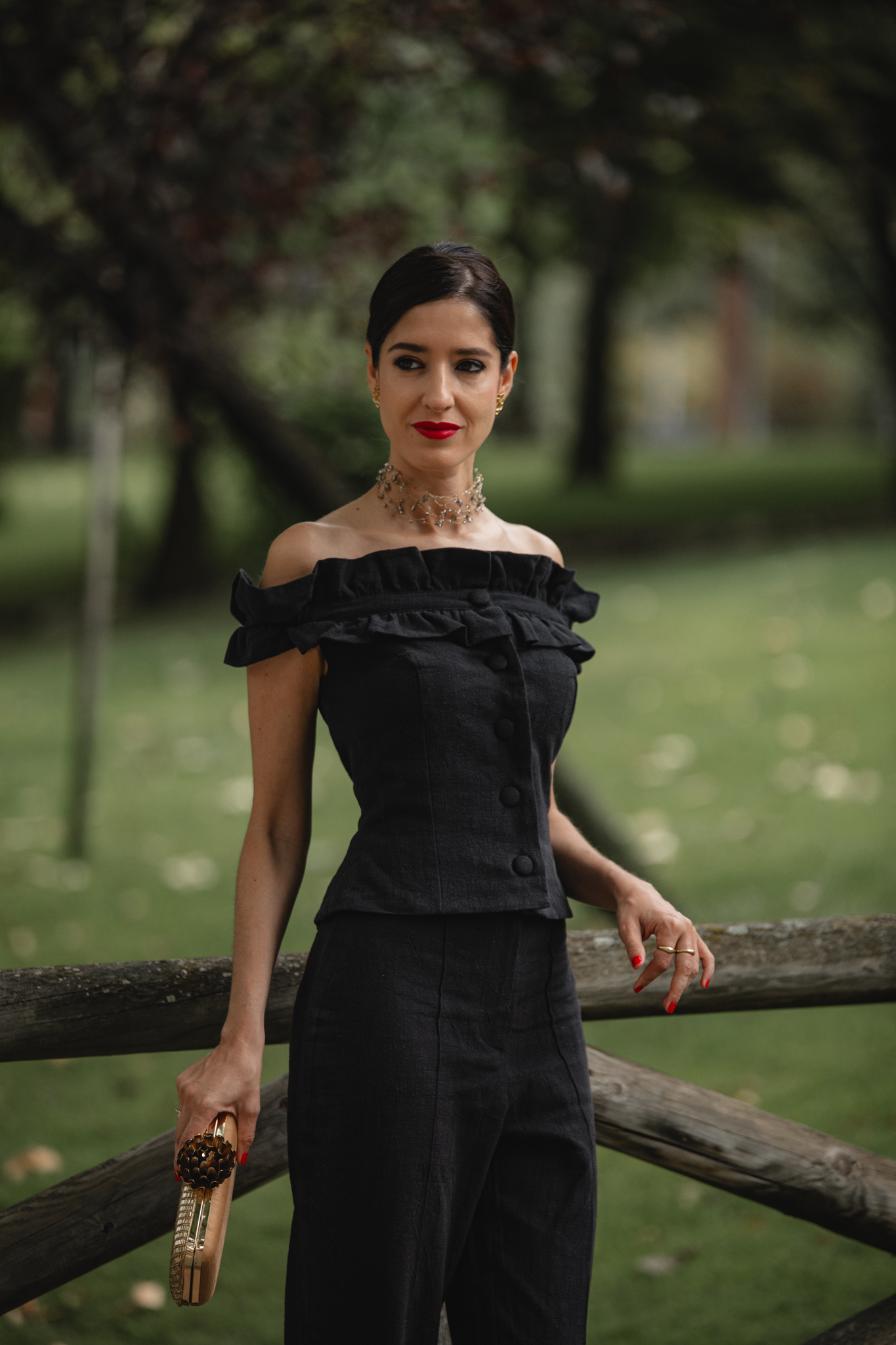 Invitada con conjunto pantalón negro de Cherubina y escote palabra de honor con volante, apoyada en una valla de madera