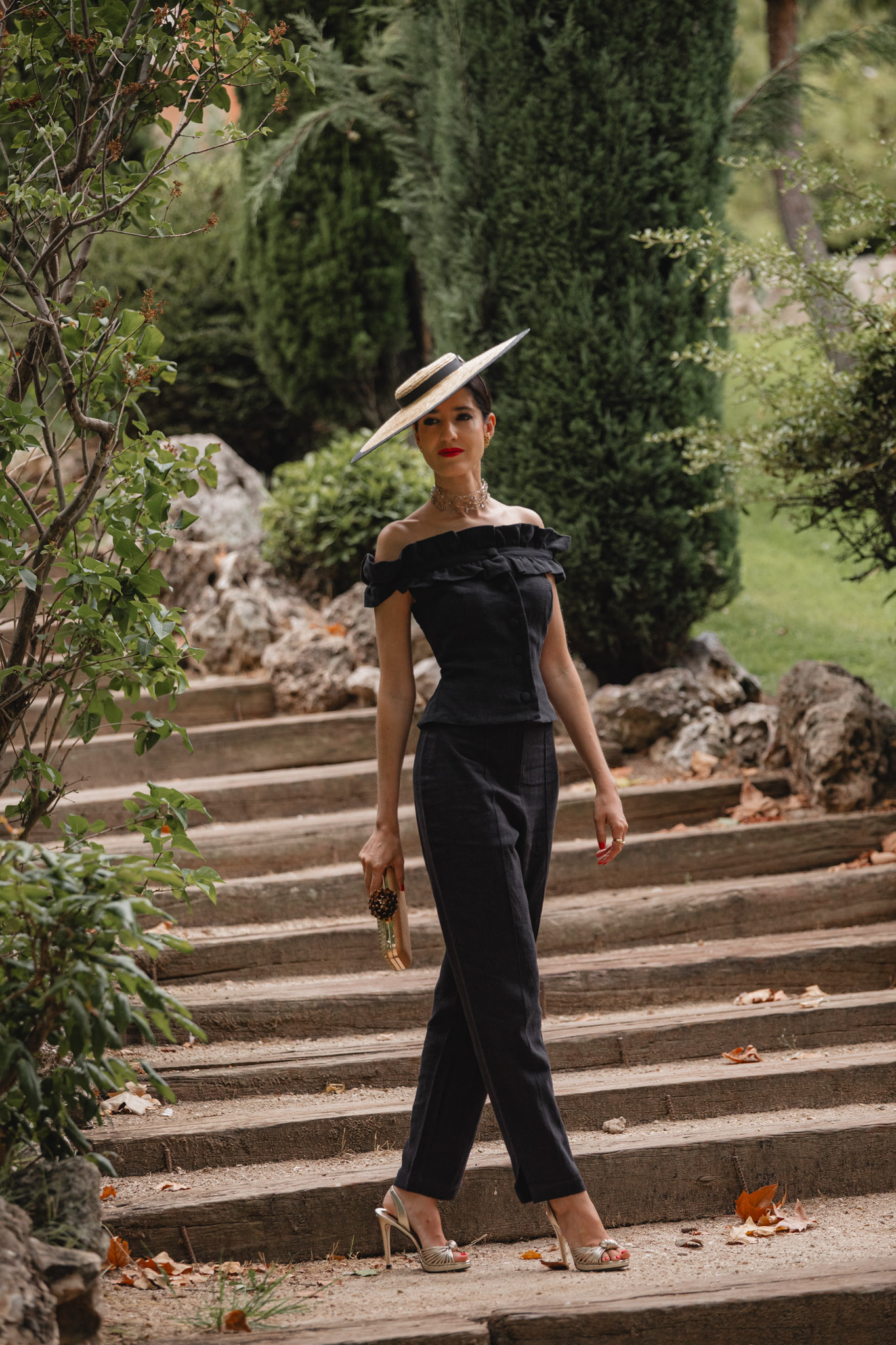 Invitada caminando por una escalinata de piedra con pamela de rafia y conjunto pantalón negro