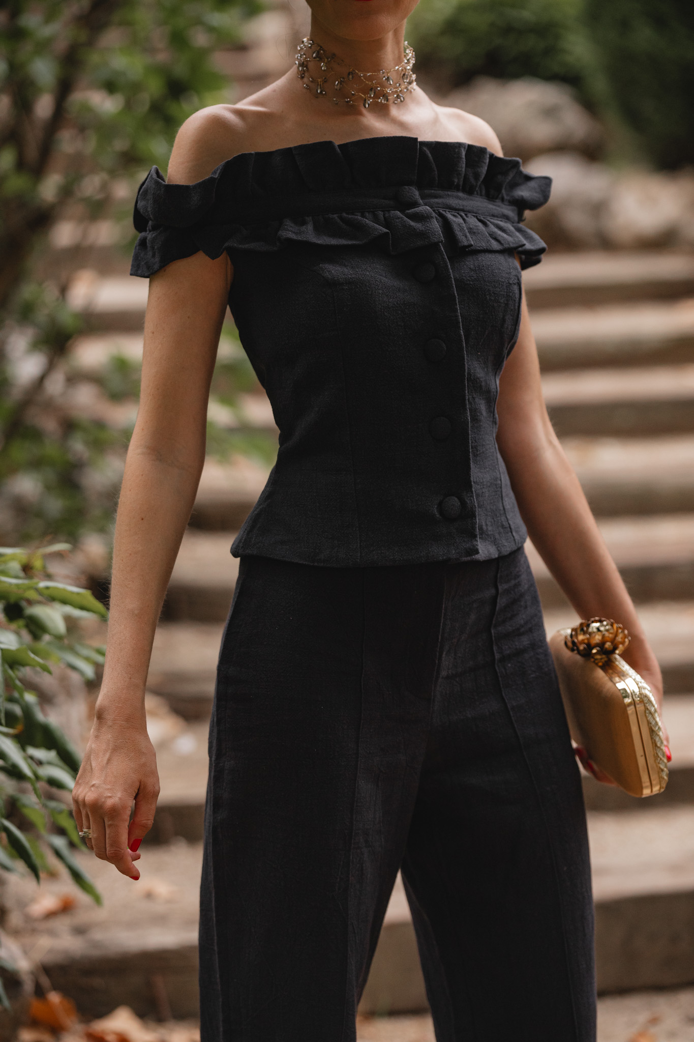 Detalle del conjunto pantalón negro de Cherubina con clutch dorado, en un jardín con escalinata de piedra