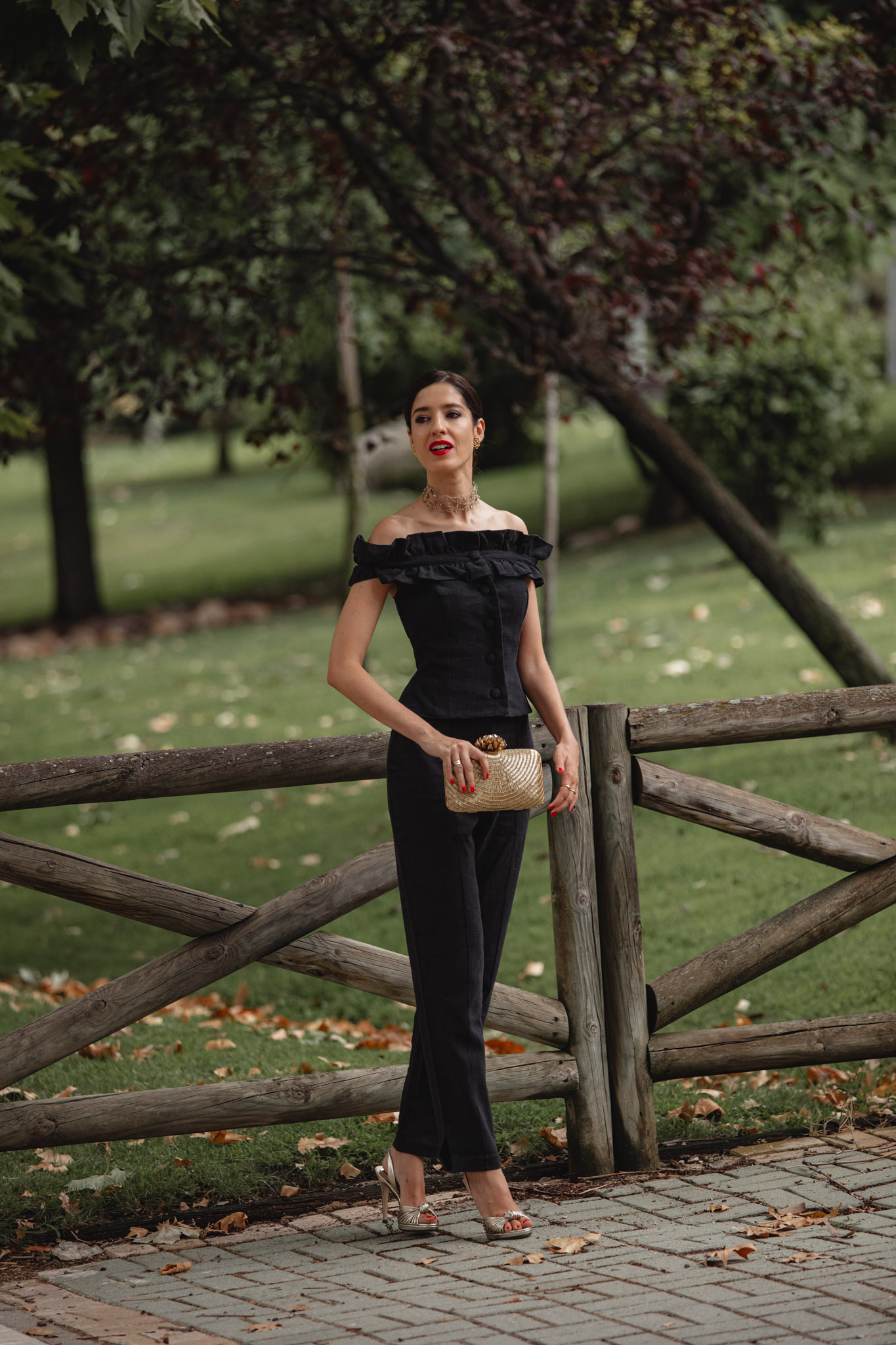 Invitada apoyada en una valla de madera con el conjunto pantalón negro y clutch dorado trenzado