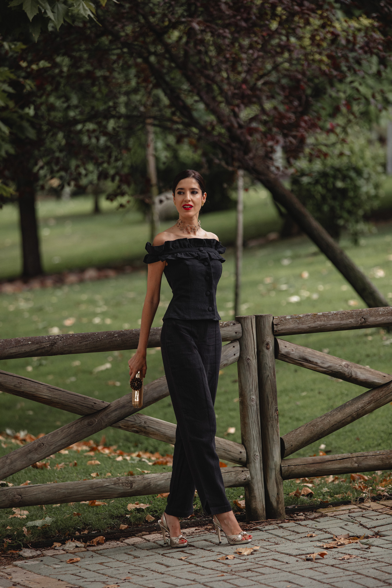 Invitada de pie junto a una valla de madera con el conjunto pantalón negro de Cherubina y clutch dorado