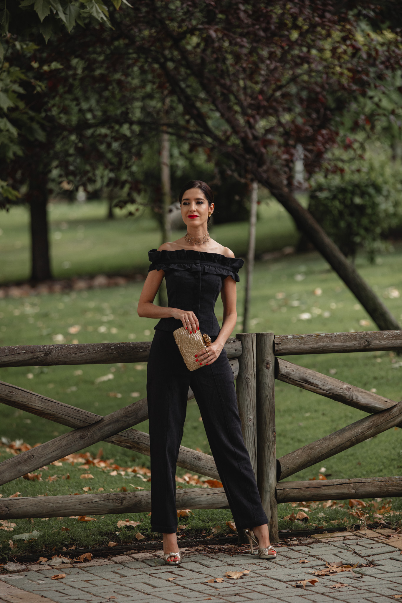 Invitada de pie junto a una valla de madera con el conjunto pantalón de Cherubina y clutch dorado
